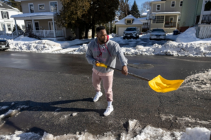 A person is shoveling snow with a yellow shovel from a paved area into a white bucket next to a red traffic cone, with a parked car and houses in the snowy background.