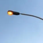 A solitary street light with a bright yellow lamp glowing against a clear blue sky.