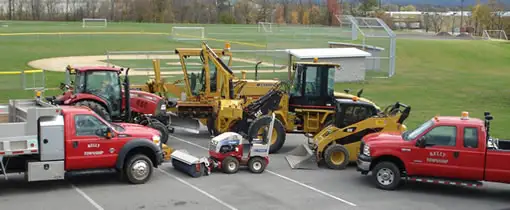Three red pickup trucks and two yellow construction vehicles, including an excavator and a bulldozer, are parked on a dirt lot in front of a white building.
