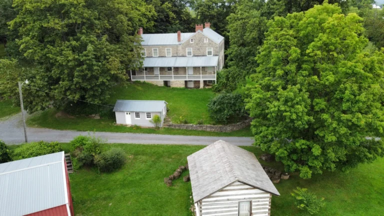 A large stone house with a wraparound porch and a smaller white outbuilding are nestled among green trees and a lawn. In the foreground, a gray-roofed wooden shed is visible, with hints of a red-roofed structure to the left.