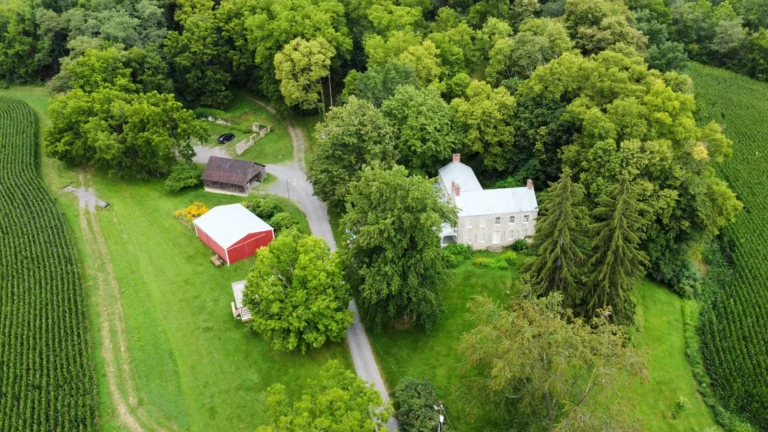 A gray stone house with a wraparound porch and a smaller white building are surrounded by green trees and a lawn. In the foreground, a gray-roofed wooden shed is visible, with a hint of a red-roofed structure to the left.