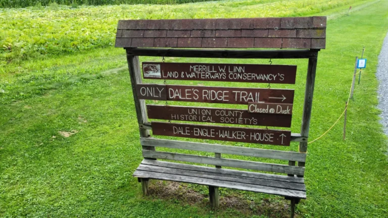 A large wooden sign with a pointed roof and a bench beneath it stands in a grassy area. The sign displays information about "Merrill W Linn Land & Waterways Conservancy," "Only Dales Ridge Trail," "Union County Historical Society," and "Dale Engle-Walker House," with arrows indicating directions.